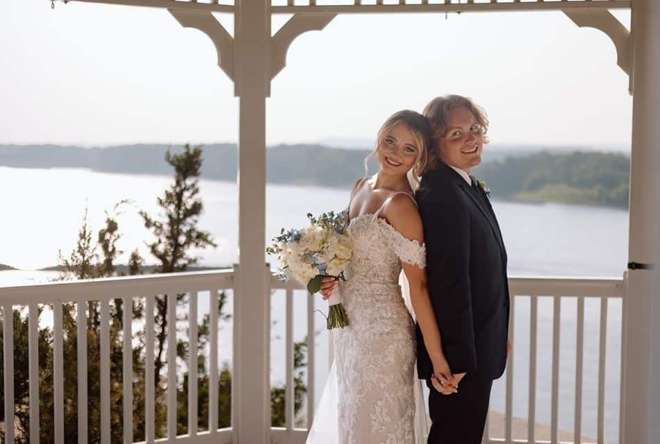 Lady in white dress holding white flowers and man in dark suit standing back to back holding hands with rail and river in background