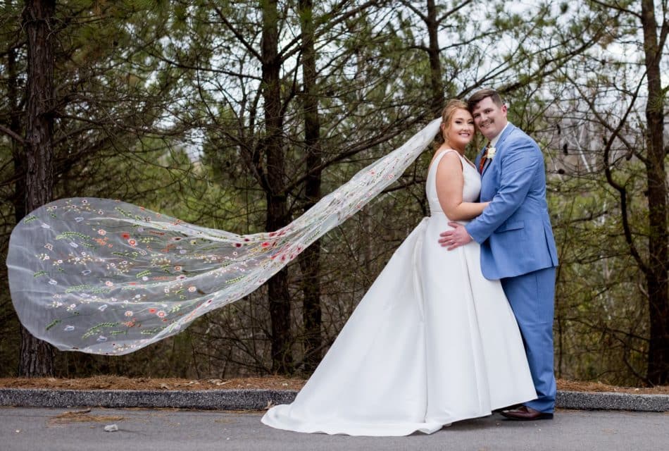 Lady in white dress with a hand-stitched veil caught by the wind with a man in a light blue suit in front of trees.