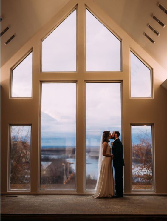 Bride and groom facing each other and kissing. They are in a chapel with a high ceiling in front of floor to ceiling windows with the river in the distance through the window.