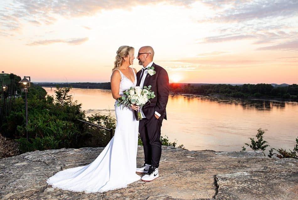 Man in black with white Nike hightops looking at a lady in a white sleeveless dress holding a bouquet of roses. They are standing on a river bluff overlooking a river with a sunset in the sky.
