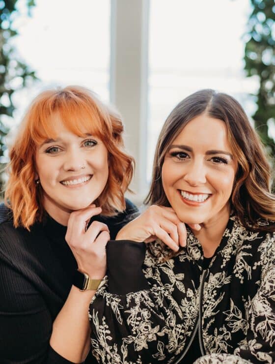 two women smiling with their hands under their chins posing together in front of large windows.