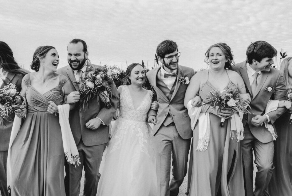 black and white photo for bride and groom locking arms with their bridal party. there are two groomsman and two bridesmaids in the picture and a groomsman and a bridesmaid who are are only partially in the pictures. They are all smiling. the bride and bridesmaids are carrying flowers.