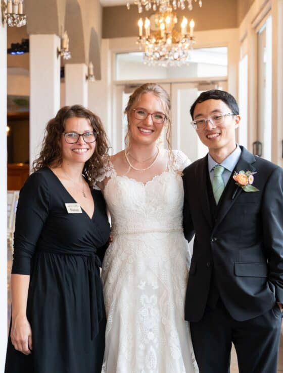 two women smiling with their hands under their chins posing together in front of large windows.