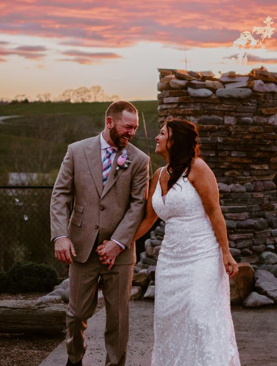 Man in tan suit holding hands of woman in white dress standing with a tall stone fireplace in the background