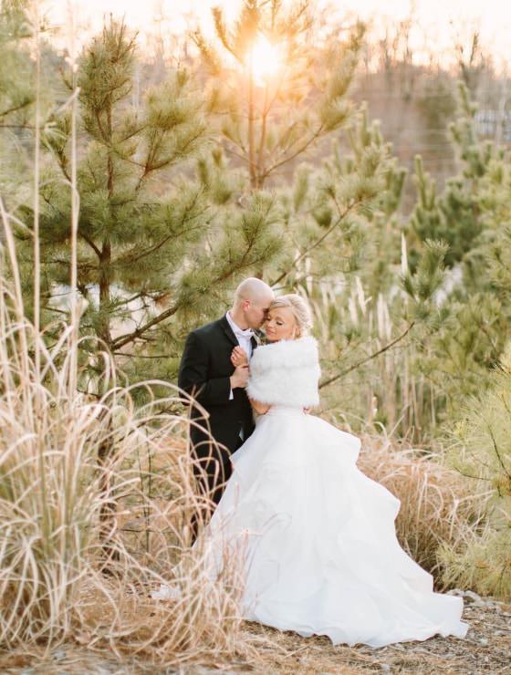 Man in black suit holding woman in white dress with evergreen trees and setting sun in the background