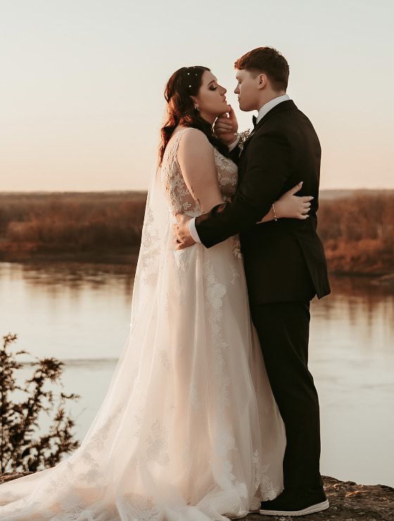 Man in black suit holding woman in white dress standing on a rock with a river and setting sun in the background