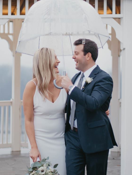 Man in gray suit holding an umbrella with other arm around woman in white dress standing near a white gazebo