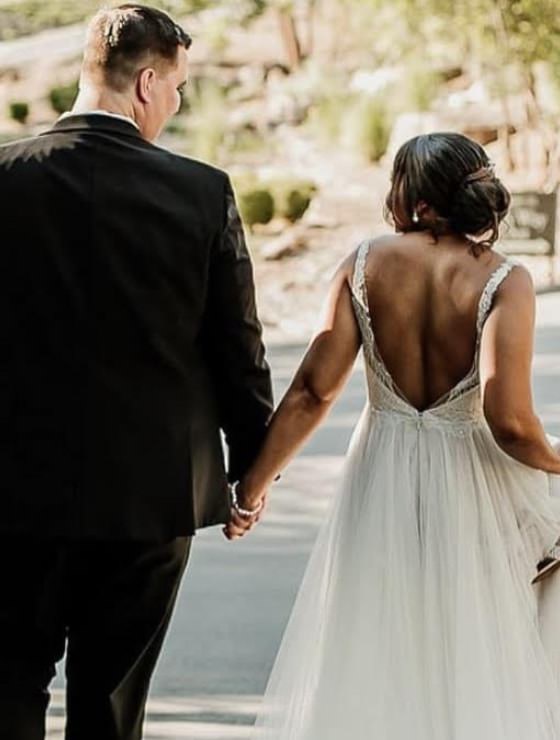 Man in black suit holding the hand of woman in white dress who is holding her shoes while they walk