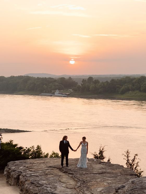 Man in black suit holding hand of woman in white dress standing on a rock with a river and setting sun in the background