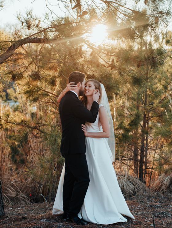 Man in black suit with arms around woman in white dress standing in front of evergreen trees with setting sun behind