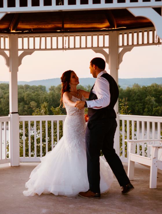 Man in black suit dancing with woman in white dress under white gazebo