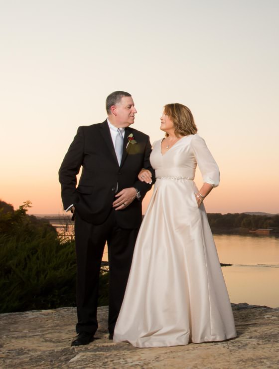 Man in black suit holding the arm of a woman in an off-white dress standing on a rock with a river and setting sun in the background