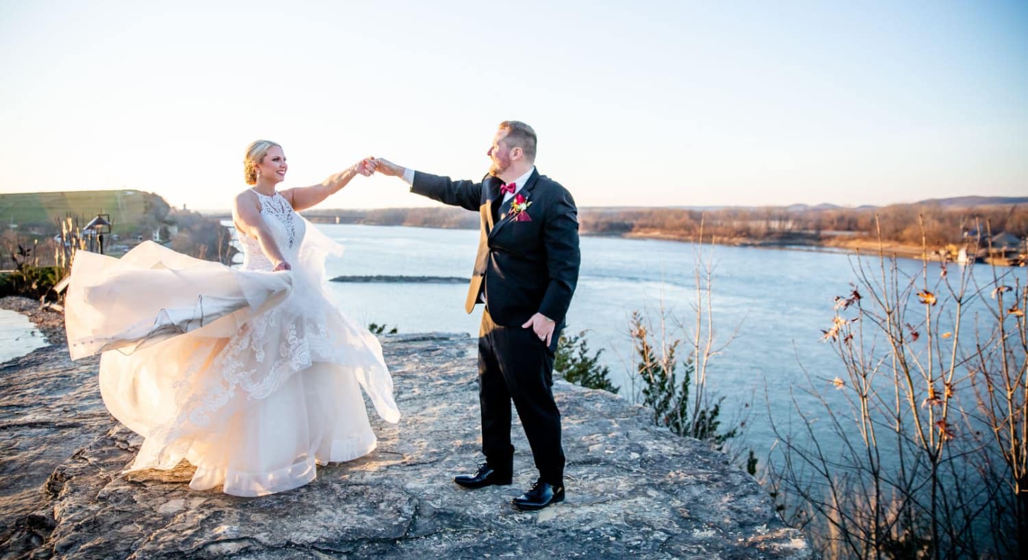 Lady in a white dress holding the hand of a man in black suit standing on a rock with a river in the background