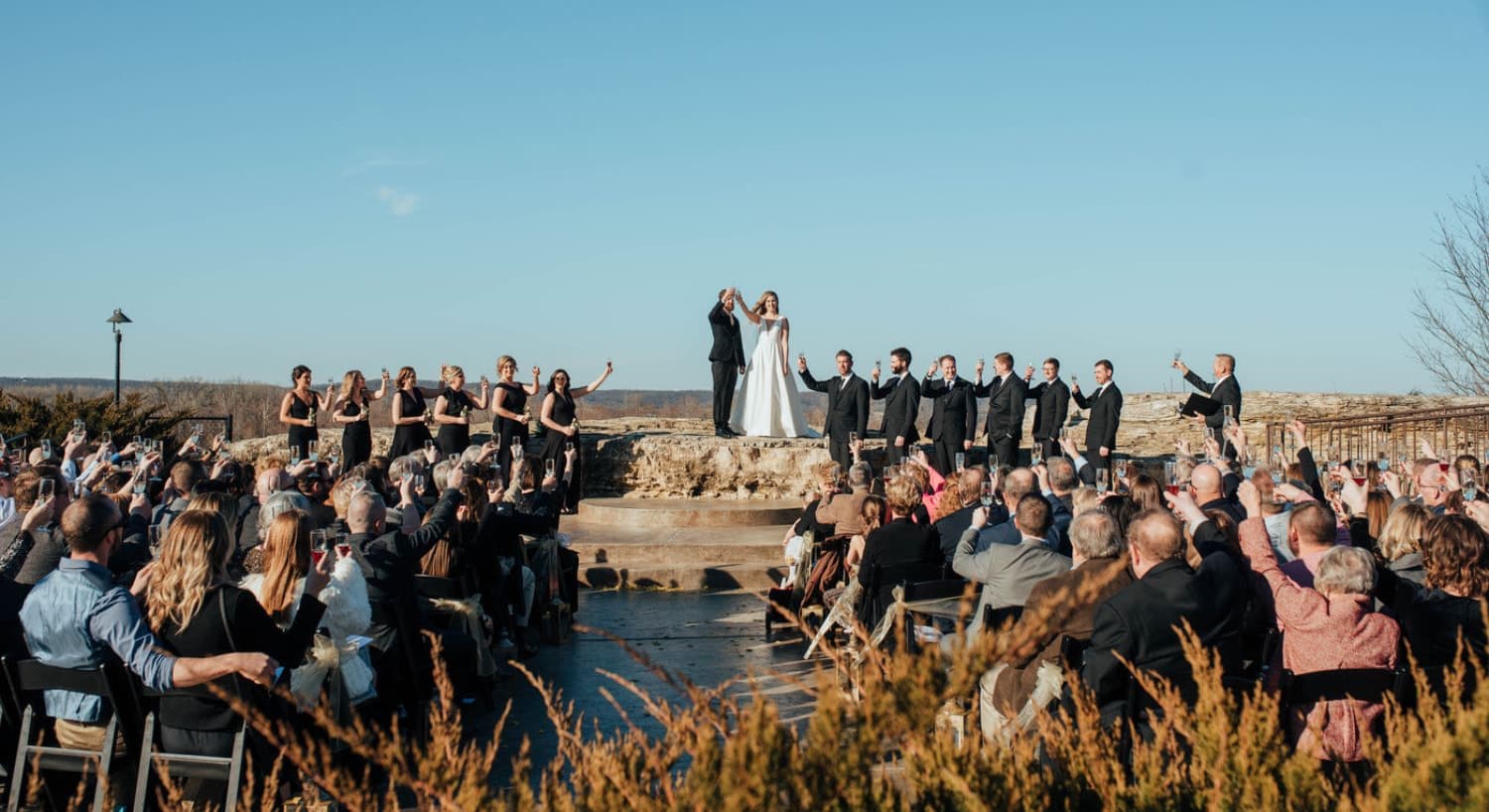 Wedding party standing on and near a large rock with seated guests all toasting the bride and groom in an outside venue