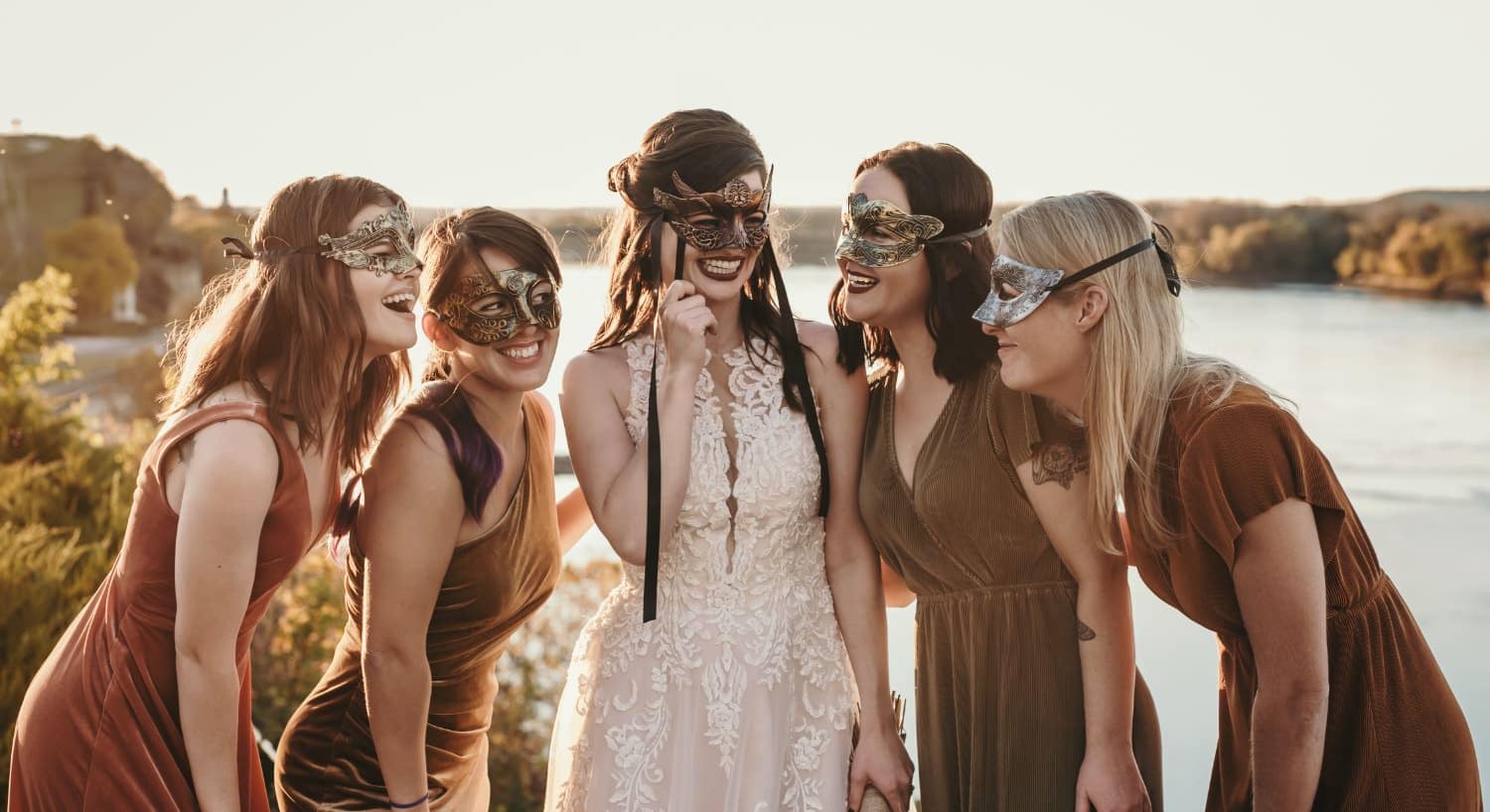 Lady in white dress and other ladies in fall color dresses wearing masquerade masks with a river in the background