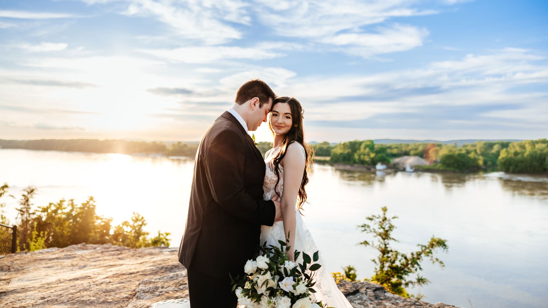 Man in black suit holding woman in white dress standing on a rock with a river and setting sun in the background