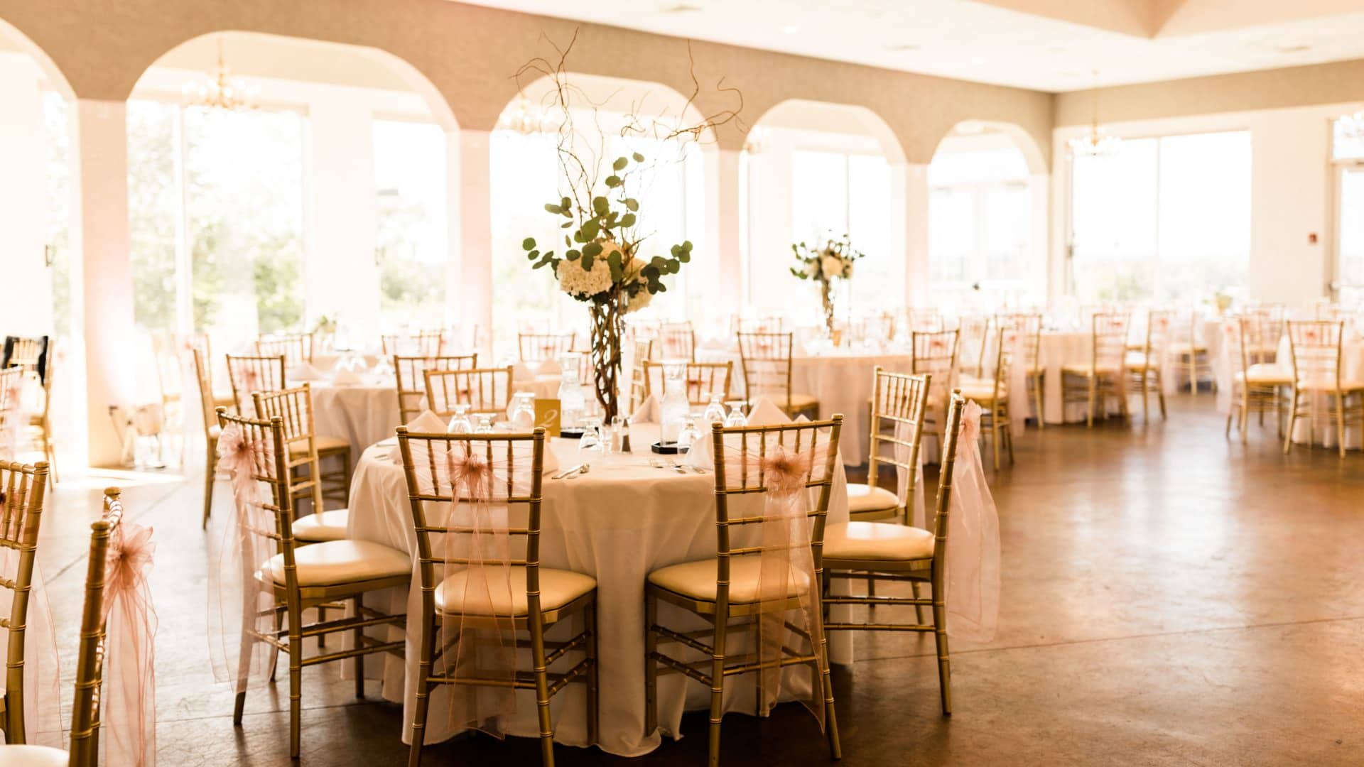 Large reception hall set up with tables with white linens, gold chairs, and large bouquets of flowers