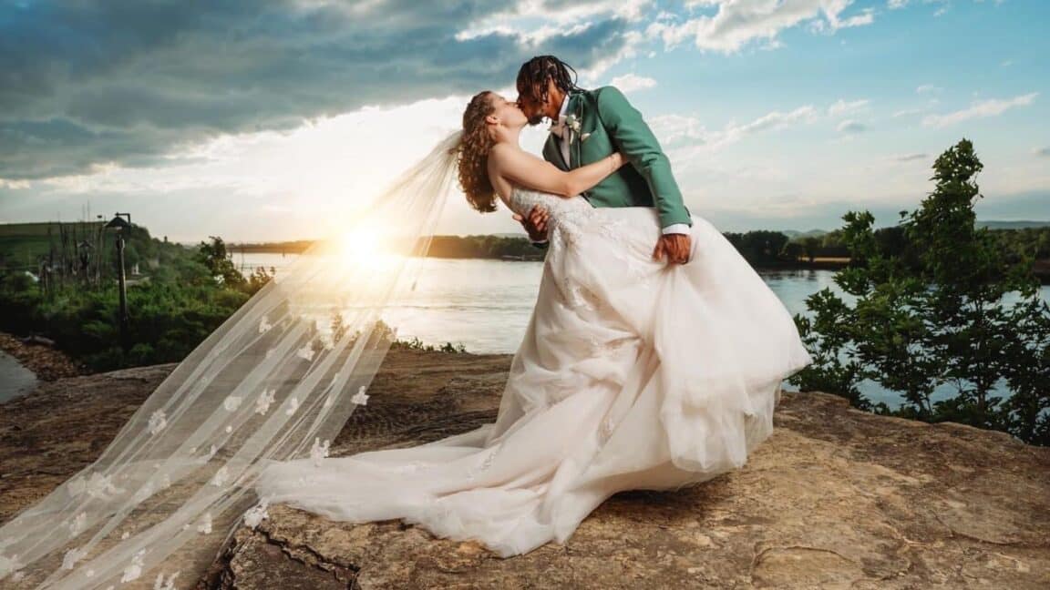 bride with a long veil and groom in a teal blue suit kissing on top of a bluff in front of a sunset over the water