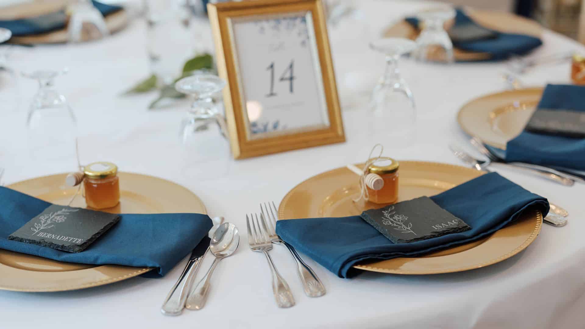 wedding table setting with white tablecloth, gold chargers, dark blue napkins, silverware, glassware, party favors, and decor