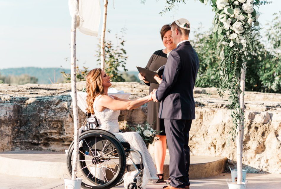 Lady in white dress sitting in wheelchair holding hands with man in black suit with stone wall in background