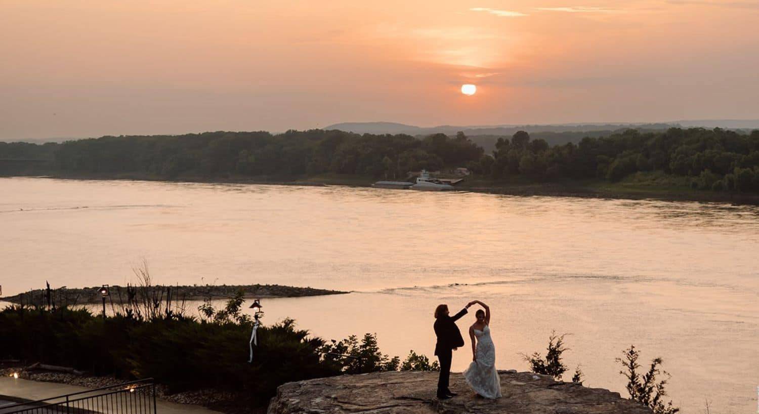 bride and groom dancing on a large flat rock over looking a river in background at sunset. Brilliant orange sunset.