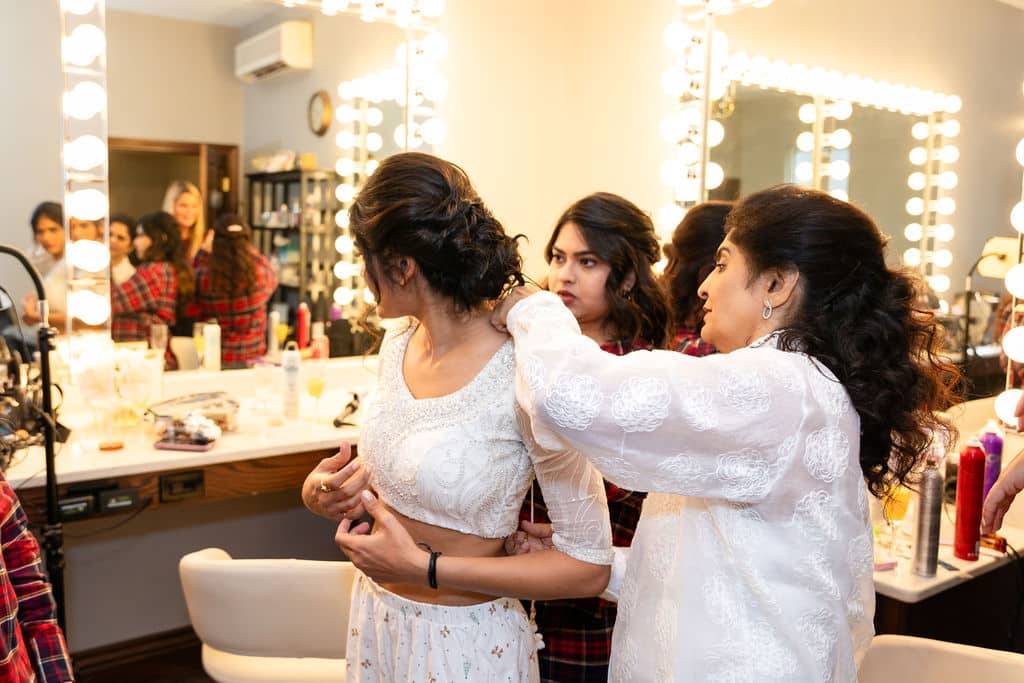 mother and bridesmaid helping bride with her hair in front of two large makeup mirrors. 