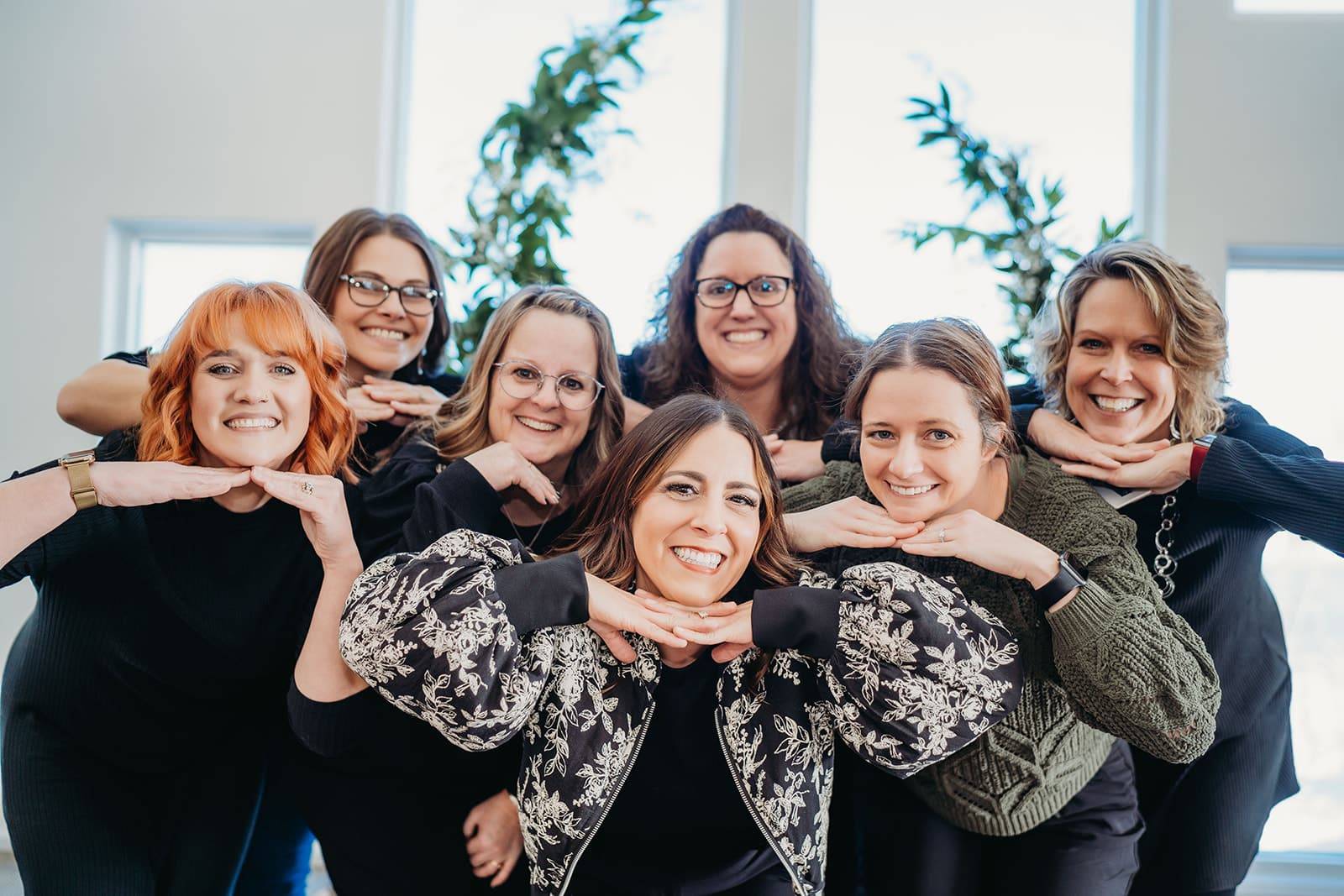 seven wedding coordinators grouped together leaning in close to one another with hands folded under chin with smiles having fun. In front of large windows. 