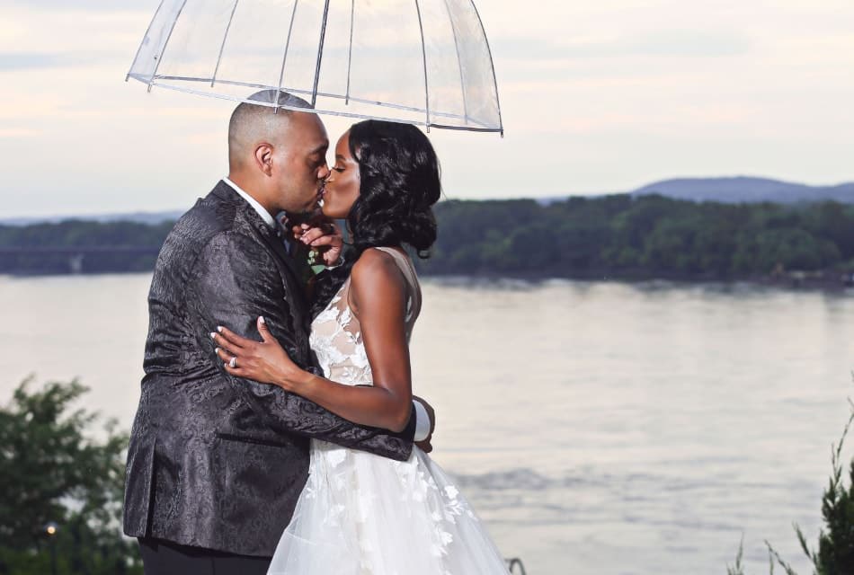 Man in dark gray suit holding umbrella and kissing lady in white dress with river in the background